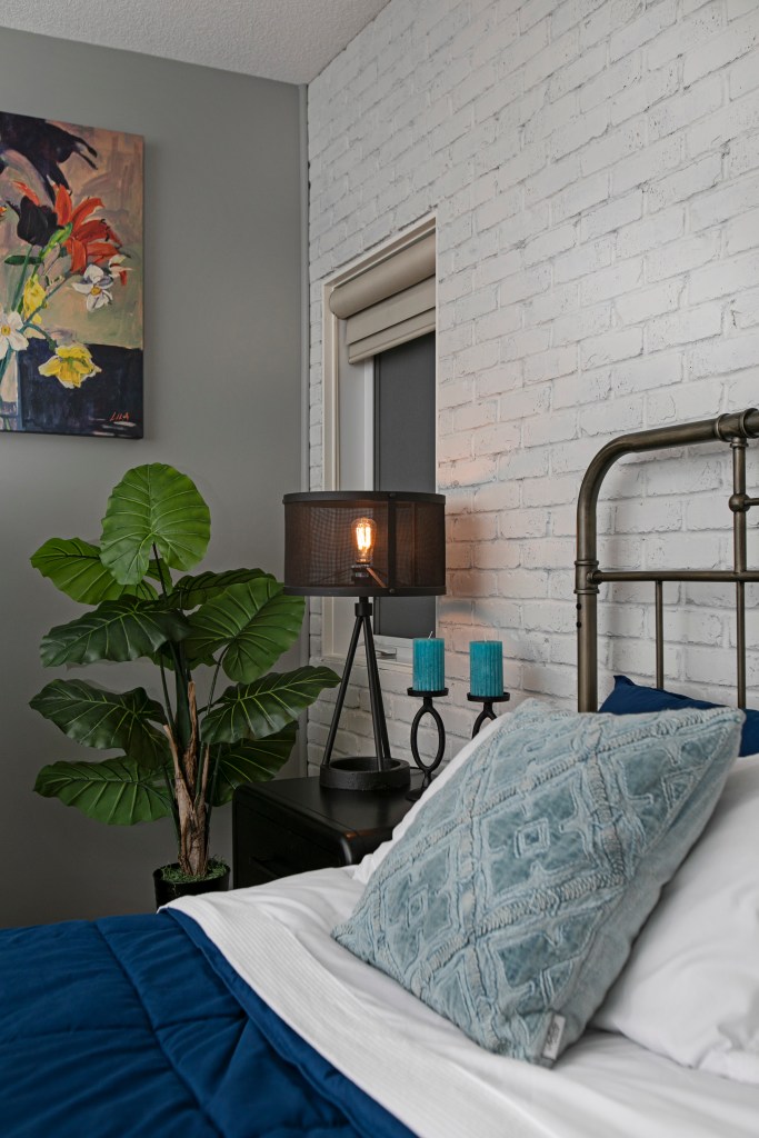 A view of an inside corner of a primary bedroom with a window beside the nightstand and a white washed brick accent wall located behind the headboard on the bed. This was part of a Ensuite bathroom renovation located in Edmonton, that was completed by Awlridge Renovations.